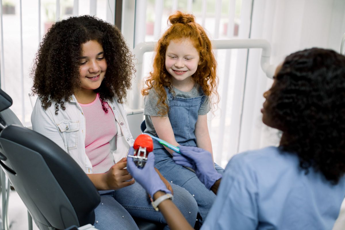 Dental hygienist demonstrates a tooth-brushing tool and dental model to two smiling girls in a clinic setting.