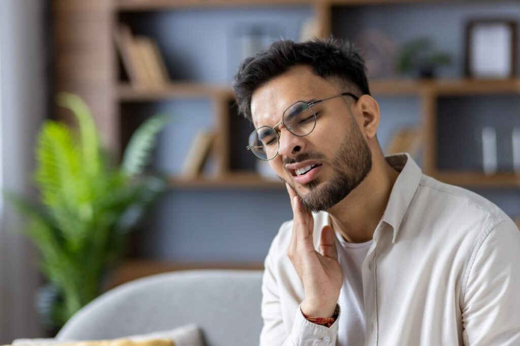 Man with round glasses sits on a sofa, touching his jaw with a pained expression in a cozy living room.