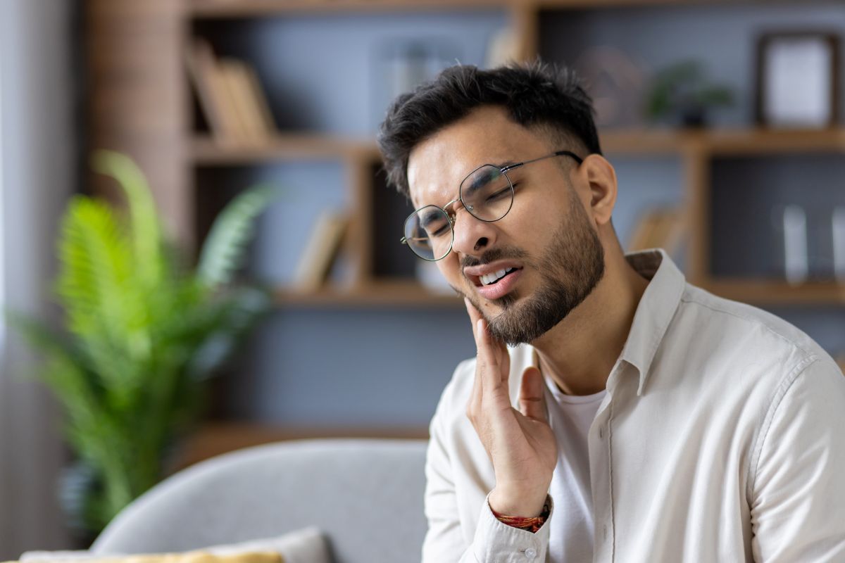 Man with round glasses sits on a sofa, touching his jaw with a pained expression in a cozy living room.