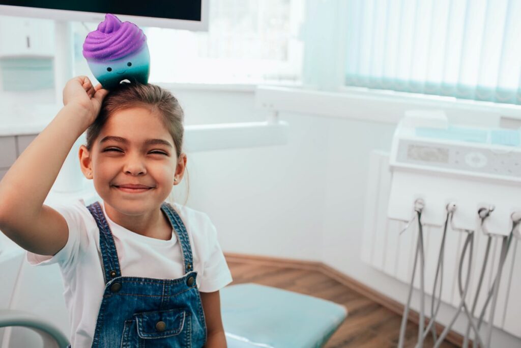 Smiling young girl in a dental clinic balancing a colorful plush cupcake on her head (purple and teal).