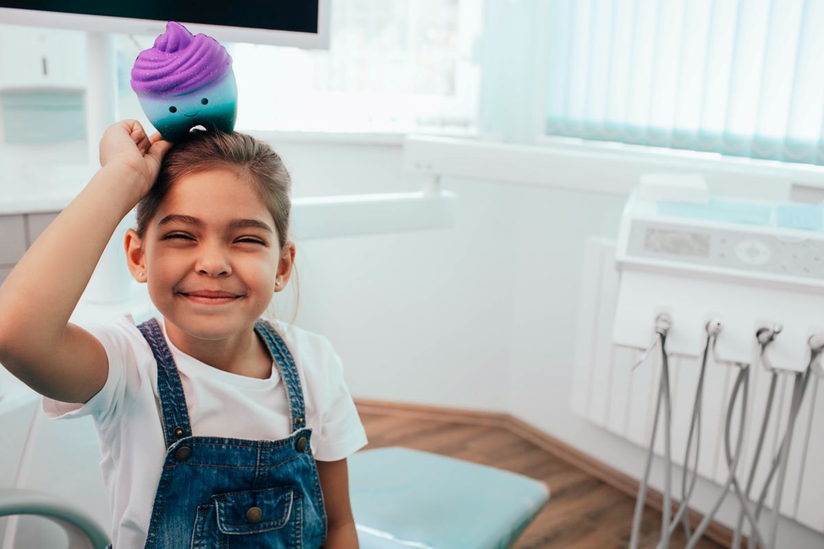 Smiling young girl in a dental clinic balancing a colorful plush cupcake on her head (purple and teal).