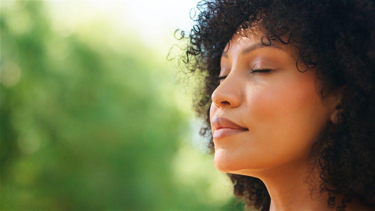 Woman with curly hair, eyes closed, savoring sunlight outdoors with a green blurred background.